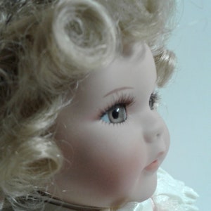 May include: A porcelain doll with blonde curly hair, wearing a white dress with pink accents. The doll has blue eyes and a painted smile.