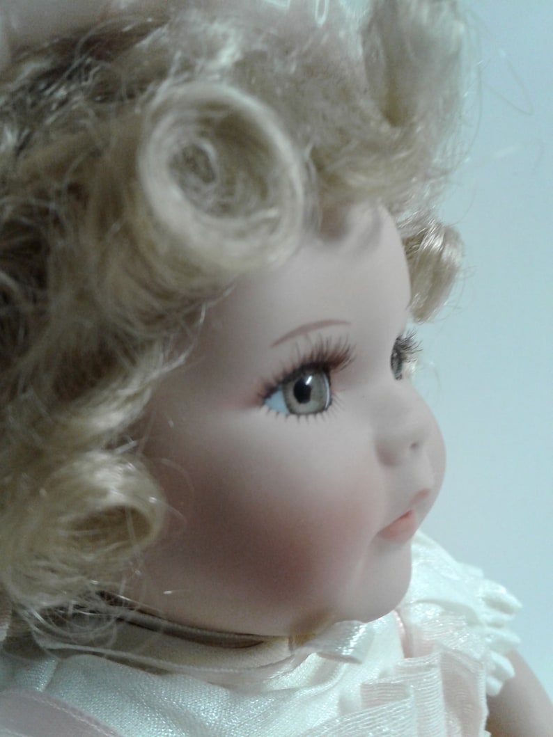 May include: A porcelain doll with blonde curly hair, wearing a white dress with pink accents. The doll has blue eyes and a painted smile.