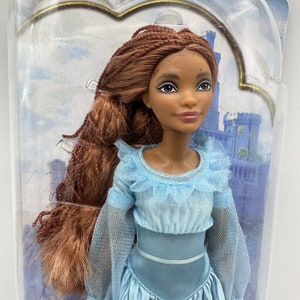 May include: A doll with brown hair styled in braids, wearing a light blue dress with a ruffled neckline. The doll has brown eyes and dark brown skin.