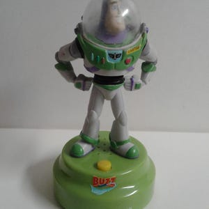 May include: A Buzz Lightyear figurine from the Toy Story franchise. The figure is wearing a green and white spacesuit with a clear helmet. It stands on a green base with a yellow button and the word "BUZZ".