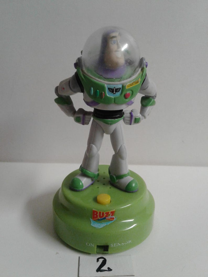 May include: A Buzz Lightyear figurine from the Toy Story franchise. The figure is wearing a green and white spacesuit with a clear helmet. It stands on a green base with a yellow button and the word "BUZZ".