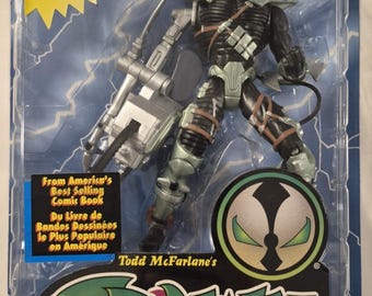 Pode incluir: Uma figura de ação Spawn The Curse da McFarlane Toys na embalagem original. A figura apresenta um capacete prateado, armadura preta e um grande braço cibernético prateado com uma motosserra. A embalagem inclui o logótipo Spawn.