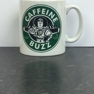 May include: White ceramic mug with a green circle design featuring the words "Caffeine Buzz" and a cartoon image of Buzz Lightyear from the Toy Story franchise.