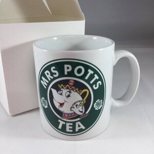 Könnte beinhalten: Weiße Keramiktasse mit einem grünen Kreisdesign, das Mrs. Potts aus Disneys Die Schöne und das Biest zeigt. Der Kreis zeigt "Mrs. Potts Tea" mit einem Cartoon-Bild von Mrs. Potts und Chip.