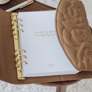 May include: A tan, quilted leather-like planner with a gold-toned six-ring binder mechanism. The white interior pages feature embossed floral patterns and the gold-printed text: Thoughtfully planned, beautifully lived. Oh So Bella my planner.