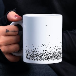 Puede incluir: Una taza de cerámica blanca con un diseño negro moteado alrededor de la parte inferior. La taza está siendo sostenida por una persona que lleva una camisa negra y pantalones vaqueros.