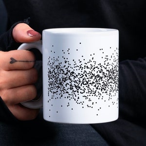 Op de afbeelding: Een witte keramische mok met een zwart-wit gespikkeld ontwerp. De mok wordt vastgehouden door een persoon die een zwart shirt en jeans draagt.