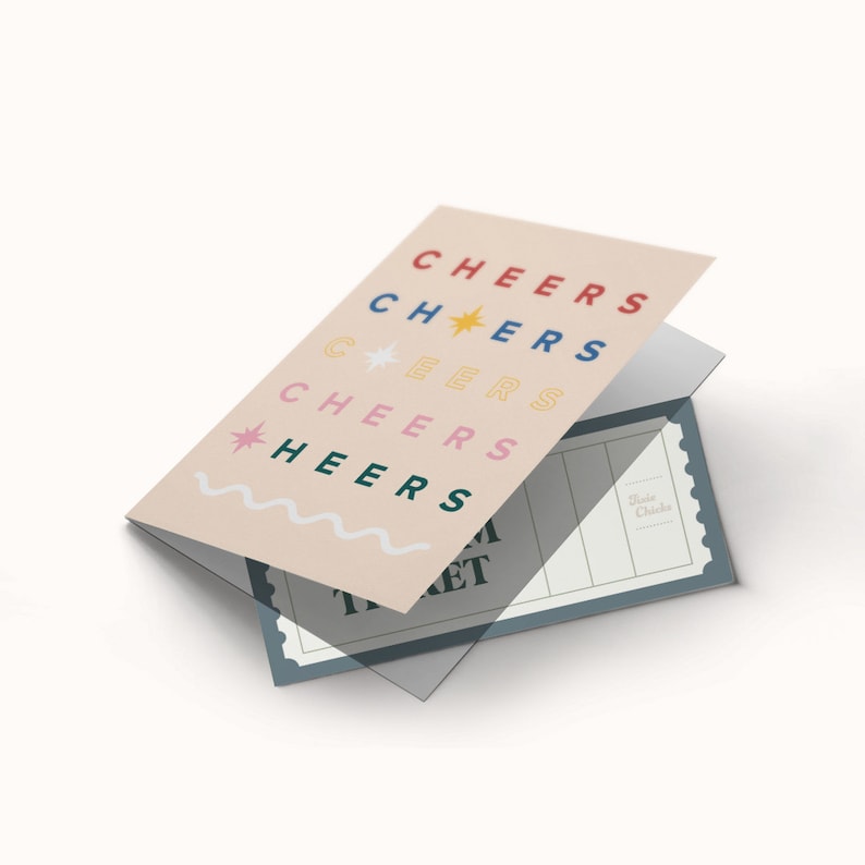 Op de afbeelding: Een roze kaart met het woord "CHEERS" vijf keer herhaald in verschillende kleuren en lettertypen. De kaart is doormidden gevouwen en eronder is een blauw ticket zichtbaar met de tekst "Ticket".