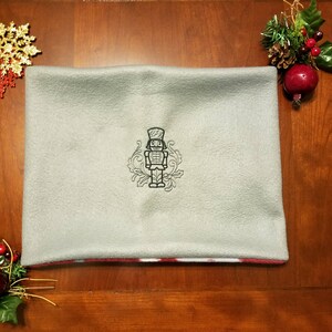 May include: A gray fleece throw pillow with a red and white trim. The pillow features an embroidered design of a nutcracker soldier.