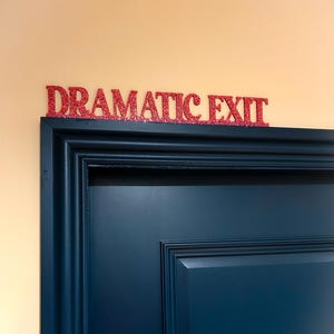 May include: A red glitter sign reading "DRAMATIC EXIT" is mounted above a dark blue door. The sign is in a serif font and is placed against a light beige wall.