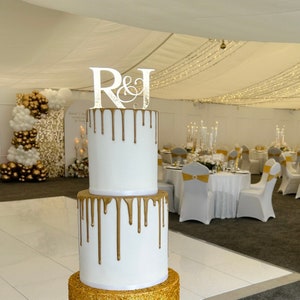 May include: A two-tier wedding cake with gold drip icing and a white fondant base. The cake is decorated with a gold glitter base and a topper that says "R&J".