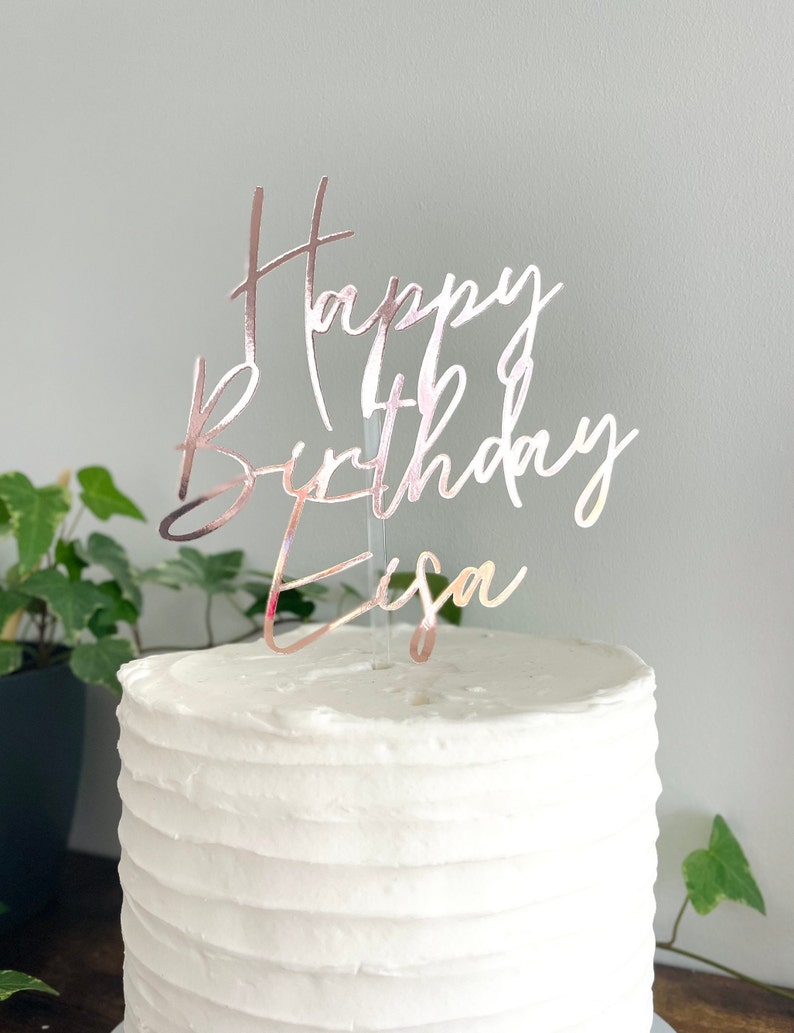 May include: A white cake with rose gold glitter cake topper that says "Happy Birthday Eisa".