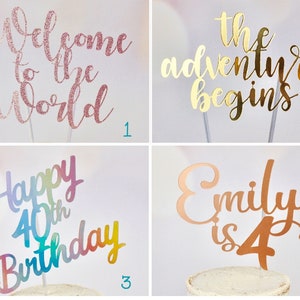 May include: Four cake toppers with different messages. The first topper is pink and says "Welcome to the World". The second topper is gold and says "The adventure begins". The third topper is rainbow coloured and says "Happy 40th Birthday". The fourth topper is copper coloured and says "Emily is 4".