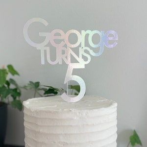 May include: A white cake with a silver iridescent cake topper that says "George turns 5".