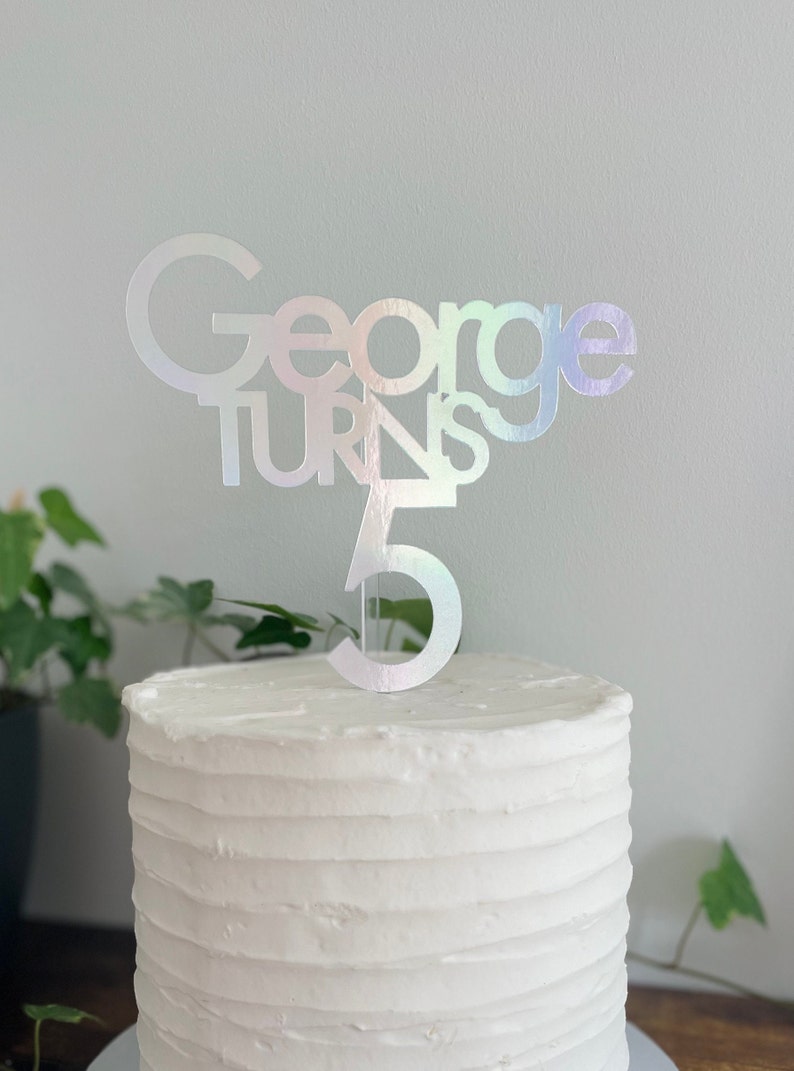 May include: A white cake with a silver iridescent cake topper that says "George turns 5".