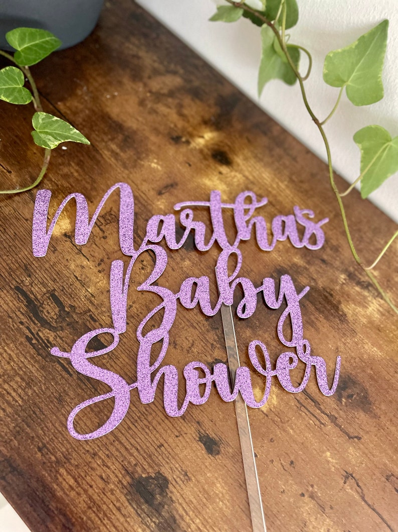 May include: Purple glitter cake topper with the text "Martha's Baby Shower" in a cursive font.