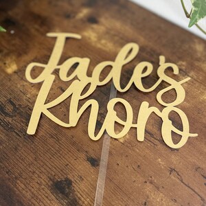 May include: A gold-colored cake topper with the text "Jades Khoro" in a cursive font.