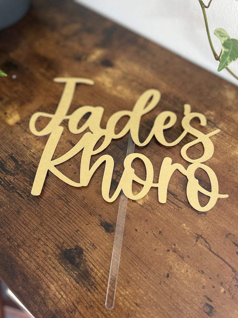 May include: A gold-colored cake topper with the text "Jades Khoro" in a cursive font.