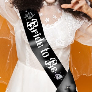 May include: A woman dressed as a bride for a Halloween party. She is wearing a white dress and veil, and has a black sash that says "Bride to Be" with a crown and spider web design.
