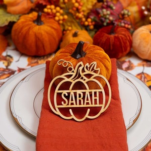 Puede incluir: Un tarjetero de madera con forma de calabaza con el nombre "SARAH" recortado. El tarjetero está sobre una servilleta naranja doblada, con un plato blanco y una calabaza de terciopelo en el fondo. También se ven otras calabazas y hojas de otoño.