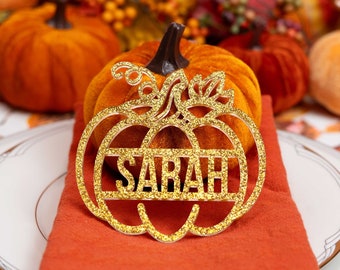 May include: A gold glitter pumpkin-shaped place card holder with the name "SARAH" cut out. The place card sits on an orange napkin, with a velvet pumpkin in the background. The table setting has a fall theme.
