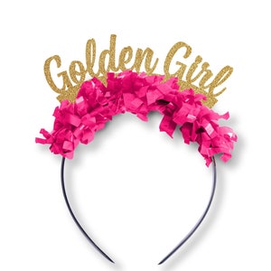 May include: A pink and gold headband with the words "Golden Girl" in gold glitter lettering. The headband is decorated with pink tissue paper.