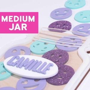 May include: A wooden jar with a white lid and a white oval label that says "Camille". The jar is filled with purple glitter, purple and blue smiley face stickers, and a few blue and purple heart stickers. The jar is labeled "MEDIUM JAR" and is part of the "Festive Gal" brand.