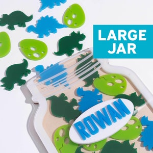 May include: A white jar with a blue and white label that says "LARGE JAR" and a blue and white logo that says "Festive Gal". The jar is filled with green and blue dinosaur and egg shapes. A white oval with the name "ROWAN" in blue letters is on the front of the jar.