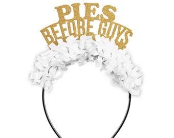 May include: A black headband with white paper fringe and a gold glitter sign that reads "PIES BEFORE GUYS". The text is in capital letters. The headband is a fun accessory for a party or event.