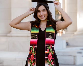 May include: A smiling person wearing a black graduation cap and dress, holding the cap. A colorful graduation stole with floral embroidery and text that reads "University of Arizona", "Class of 2025", "Biochemical Engineering", and "Victoria Espinoza".