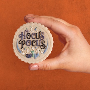May include: A round, scalloped-edge sugar cookie featuring the text Hocus Pocus in dark purple and black glittery icing. Below the text are three small potion bottles in blue, green, and purple, accented with white stars and a crescent moon.