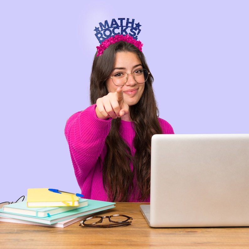 May include: A young person wearing glasses and a pink sweater with a "MATH ROCKS" headband is sitting at a desk with a laptop and school supplies. They are pointing at the camera with their right hand.