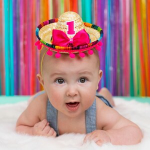 Op de afbeelding: Een baby draagt een kleurrijke sombrero met een nummer &eacute;&eacute;n erop. De sombrero is gemaakt van stro en heeft roze pompoenen rond de rand. De baby draagt een blauwe denim overall.