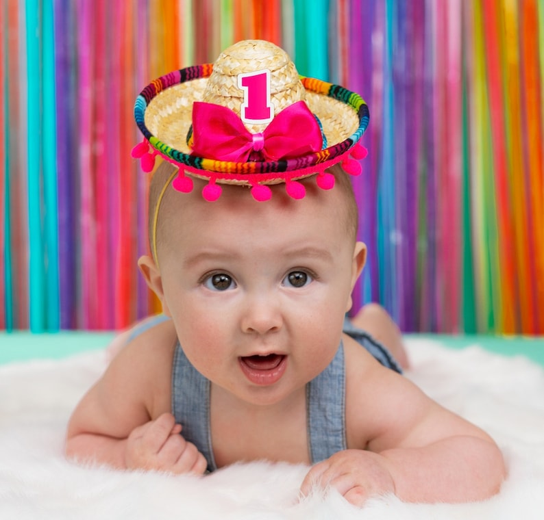 Op de afbeelding: Een baby draagt een kleurrijke sombrero met een nummer &eacute;&eacute;n erop. De sombrero is gemaakt van stro en heeft roze pompoenen rond de rand. De baby draagt een blauwe denim overall.