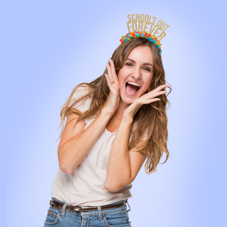 May include: A woman wearing a white t-shirt and blue jeans is smiling and holding her hands to her face. She is wearing a gold glitter headband that says "Schools Out Forever".