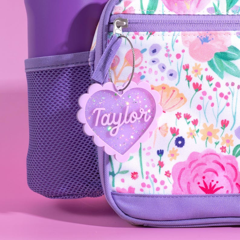 Op de afbeelding: Een gepersonaliseerde hartvormige bagagelabel met de naam "Taylor" in een sierlijk lettertype. De tag is lichtpaars met glitter en een geschulpte rand. De tag hangt aan een bloemenrugzak met een paarse mesh-zak.