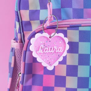 Op de afbeelding: Een roze hartvormig bagagelabel met de naam "Laura" in witte letters, bevestigd aan een rugzak. Het label heeft een geschulpte rand en is gevuld met glitter. De rugzak heeft een dambordpatroon in roze, paarse en blauwe tinten.