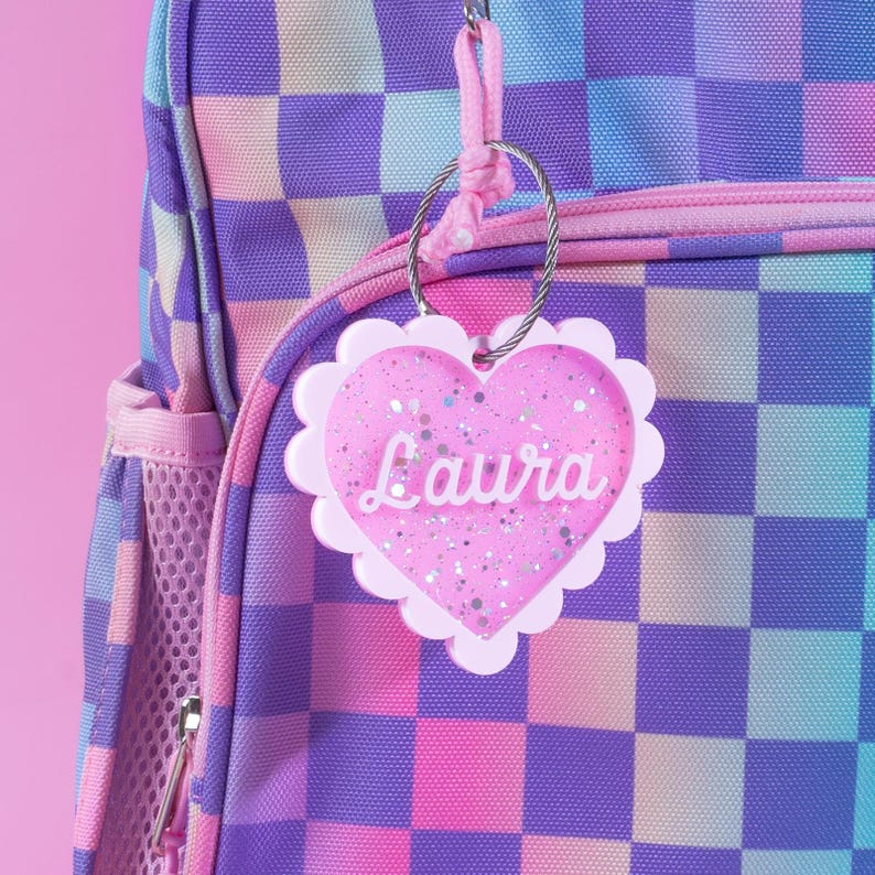 Op de afbeelding: Een roze hartvormig bagagelabel met de naam "Laura" in witte letters, bevestigd aan een rugzak. Het label heeft een geschulpte rand en is gevuld met glitter. De rugzak heeft een dambordpatroon in roze, paarse en blauwe tinten.
