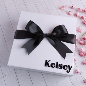May include: A white gift box with a black satin ribbon bow and the name "Kelsey" printed in black letters.