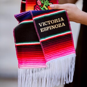 May include: A colorful, fringed graduation sash with the name "VICTORIA ESPINOZA" embroidered in gold. The sash features horizontal stripes in red, pink, black, and green, with a floral design on the top portion.