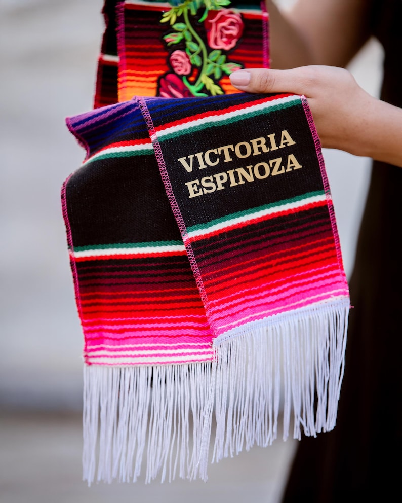 May include: A colorful, fringed graduation sash with the name "VICTORIA ESPINOZA" embroidered in gold. The sash features horizontal stripes in red, pink, black, and green, with a floral design on the top portion.