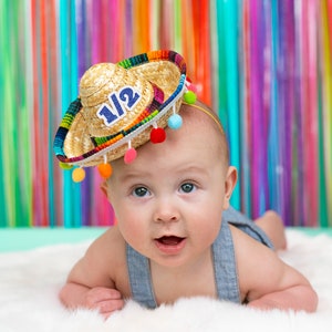 Op de afbeelding: Een baby draagt een strooien sombrero met kleurrijke pompoenen en het getal "1 1/2" erop. De baby ligt op een wit, pluche oppervlak met een kleurrijke gestreepte achtergrond.