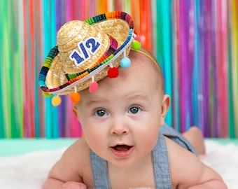 Könnte beinhalten: Ein Baby trägt einen Strohhut mit bunten Bommeln und der Zahl "1 1/2" darauf. Das Baby liegt auf einer weißen, flauschigen Oberfläche mit einem bunten gestreiften Hintergrund.