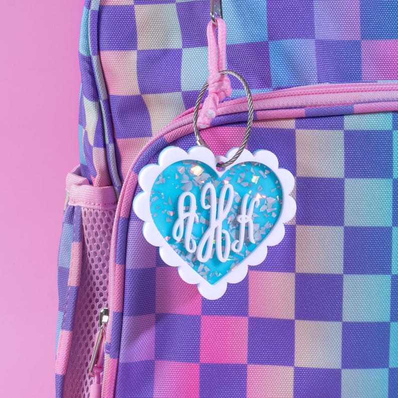 Op de afbeelding: Een hartvormig bagagelabel met een witte geschulpte rand en een blauw midden gevuld met glitter en bloemaccenten. Het label heeft de initialen "AKH" in een wit scriptlettertype, bevestigd aan een rugzak met een dambordpatroon.
