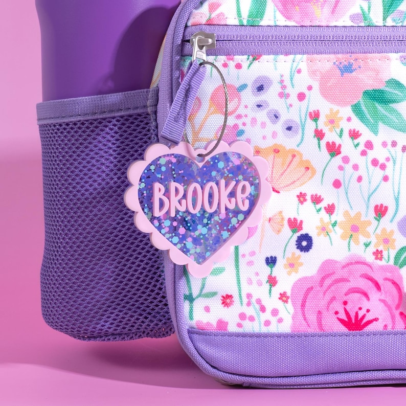 Op de afbeelding: Een roze hartvormig bagagelabel met de naam "Brooke" in witte letters. Het label heeft een geschulpte rand en is gevuld met glitter. Het label is bevestigd aan een rugzak met bloemenpatroon met een paarse rits en bies.