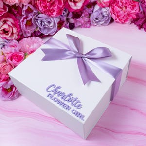May include: White gift box with a lavender ribbon and bow. The box is decorated with the text "Charlotte Flower Girl" in lavender lettering.