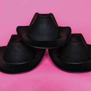 K&ouml;nnte beinhalten: Drei schwarze Cowboyh&uuml;te, die &uuml;bereinander auf einem rosa Hintergrund gestapelt sind.