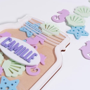 May include: A white mason jar with a clear lid filled with pastel colored seahorse, starfish, and seashell shapes. The jar has the name "Camille" written on it in purple letters.