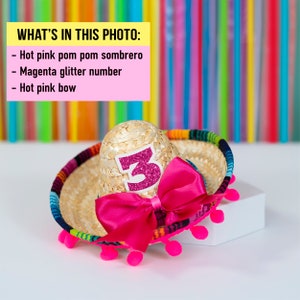 Op de afbeelding: Een kleine strooien sombrero met een roze pompon rand en een roze strik. De sombrero heeft een magenta glitter nummer "3" aan de voorkant.