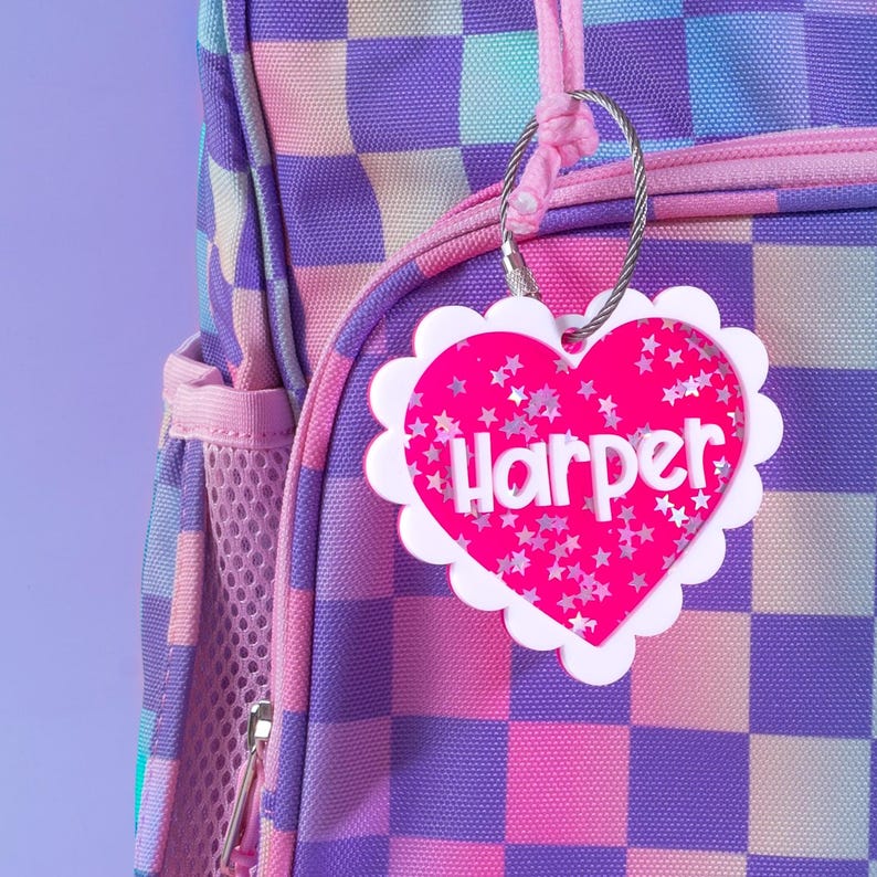 Op de afbeelding: Een roze hartvormig bagagelabel met de naam "Harper" in witte letters en sterren-glitter. Het label hangt aan een rugzak met een paars en roze geruit patroon.